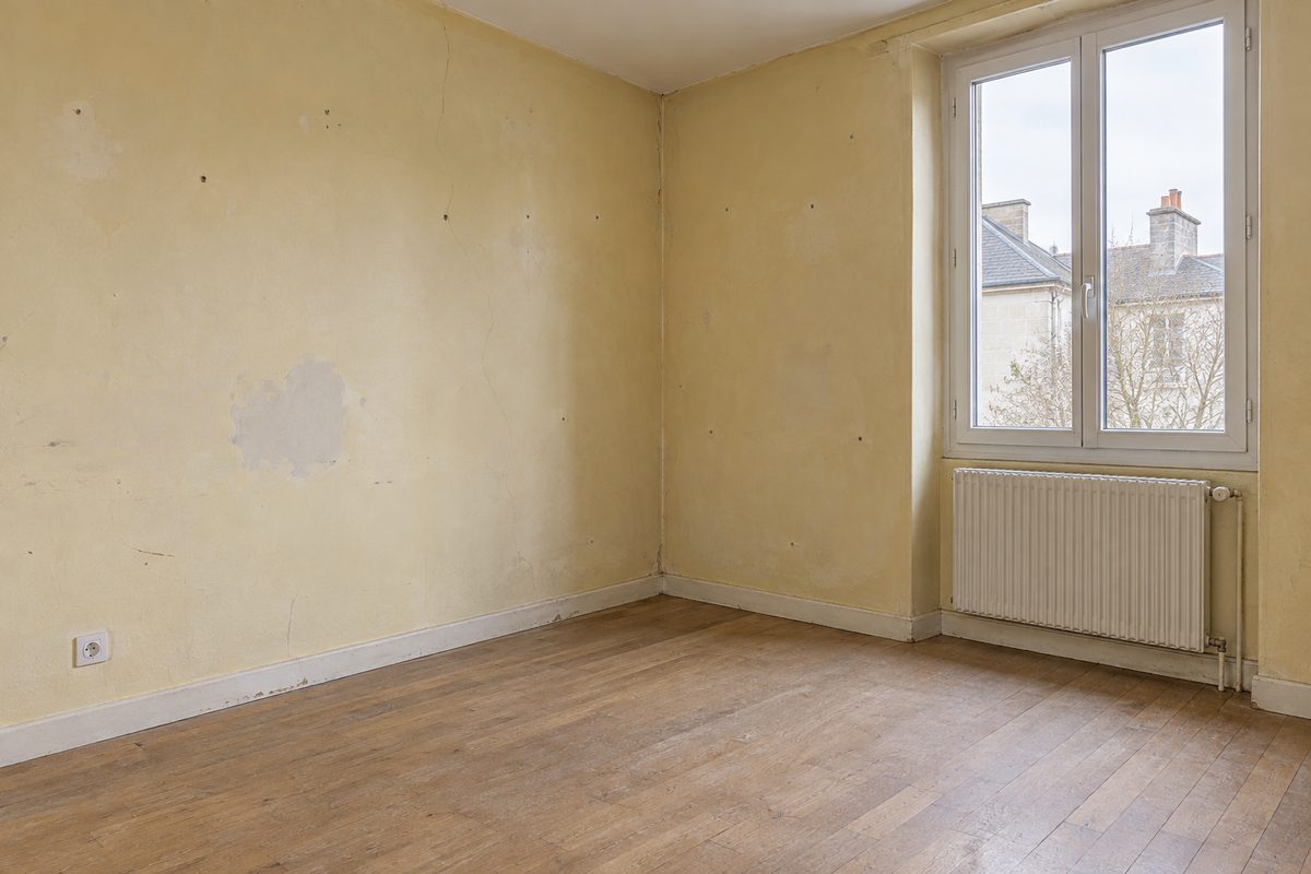 La même chambre avant travaux : murs jaunis et fissurés, plâtre écaillé, plinthes marquées et parquet usé.
